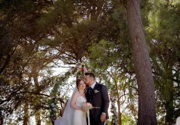 Fotografo di matrimonio in Sicilia ritrae gli sposi sotto un arco floreale nel boschetto del Baglio Trinità a Castelvetrano, mentre si scambiano un tenero gesto d'affetto dopo la cerimonia.