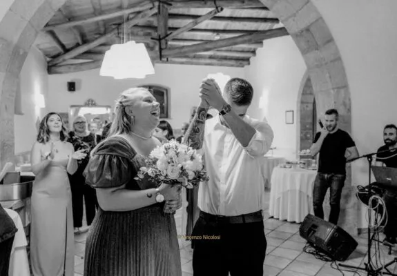 Il momento del lancio del bouquet, con la ragazza che lo riceve e il fidanzato che le prende la mano tra sorrisi, applausi e divertimento durante il ricevimento.