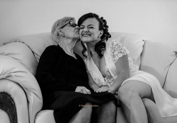 Fotografo di matrimonio in Sicilia ritrae la sposa durante la preparazione mentre riceve un affettuoso bacio dalla nonna, in un momento spontaneo e ricco di emozione.